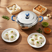 Stainless steel idli steamer with blue handles surrounded by plates of idlis, wooden steamers with dumplings and fried snacks, chutneys, ginger, and curry leaves on wooden table